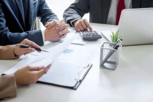 three people at a desk looking over papers with a calculator and a laptop.